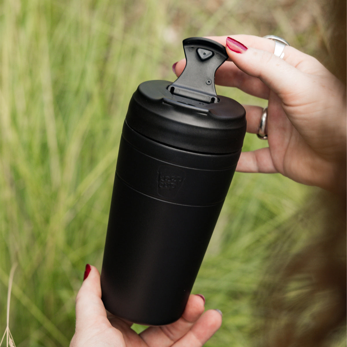 Hand opening the flip lid of a black KeepCup Commuter 454ml against a green grass background