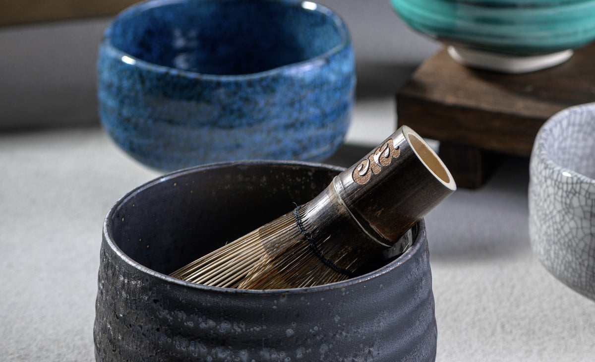Black stoneware matcha bowl with Era dark bamboo chasen, blue and teal Japanese chawan behind