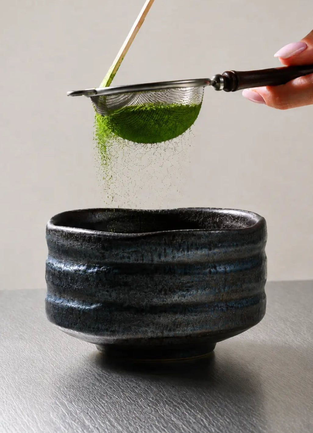 Sifting ceremonial matcha powder into a black ceramic chawan matcha bowl on a gray textured surface
